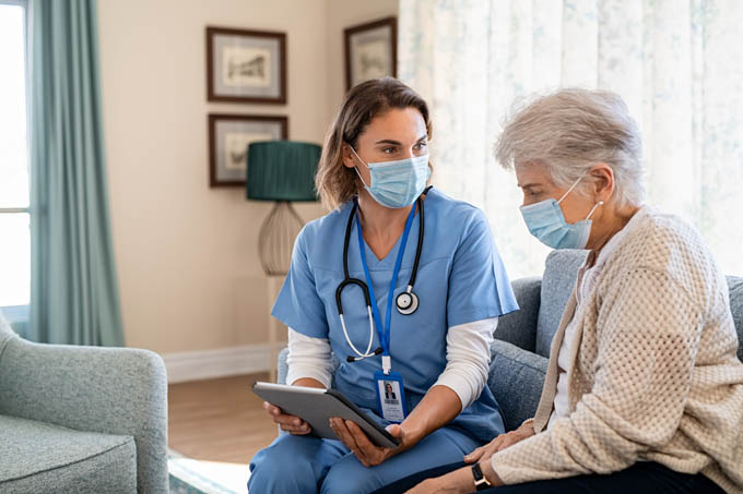 Young nurse wearing a mask and senior woman going through medical record on digital tablet during home visit