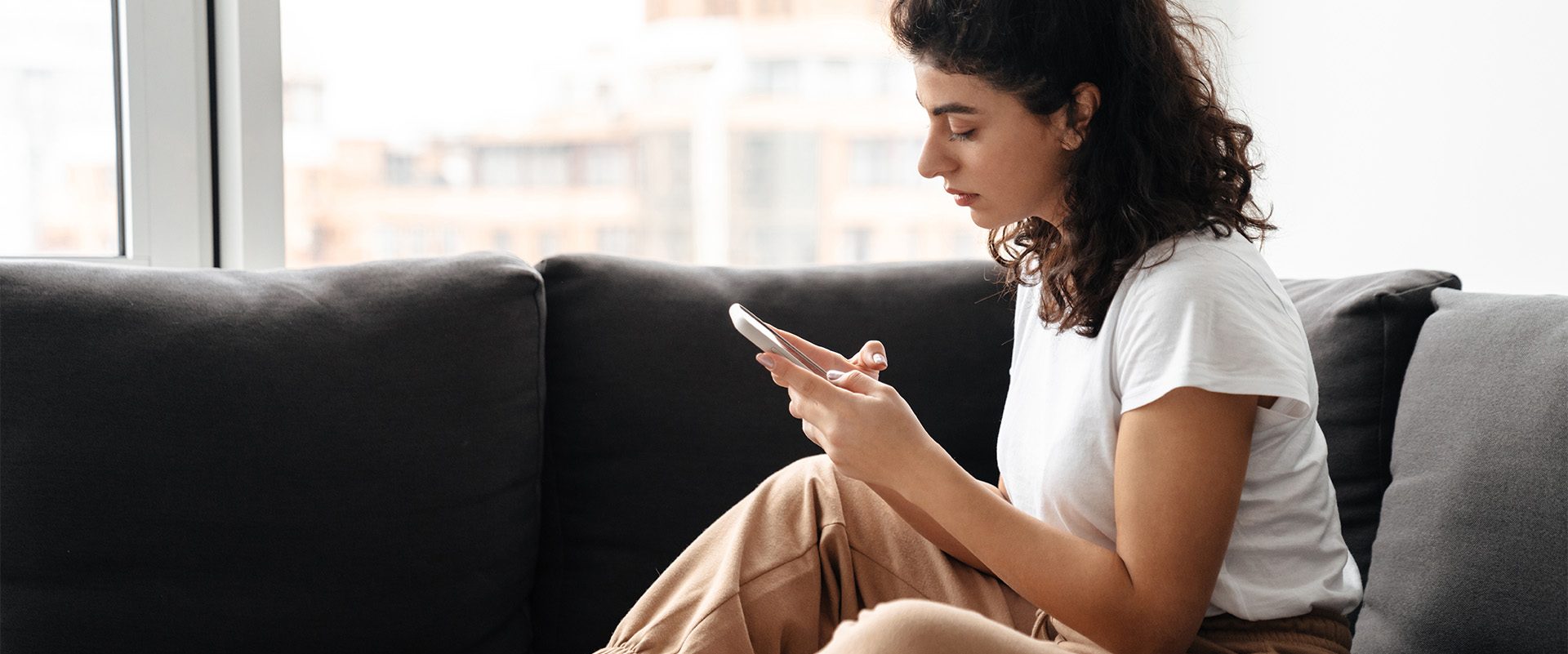 Focused brunette young woman using mobile phone while sitting on sofa